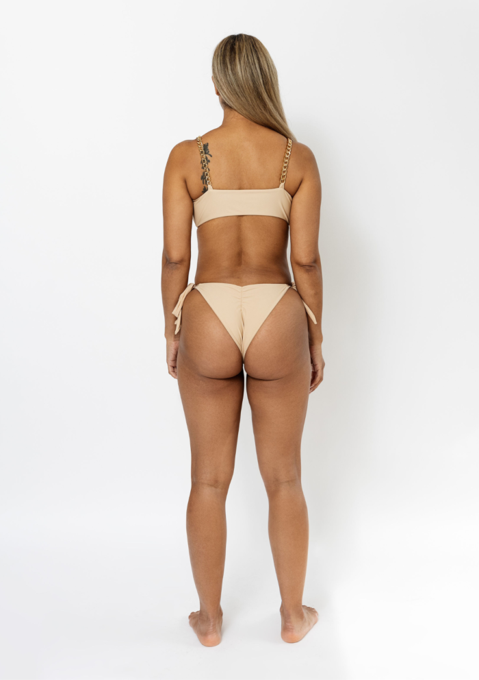 The back view of a tanned blonde model standing in an all white studio wearing a frontal tie up bikini top and matching bottoms by Vanity Couture.