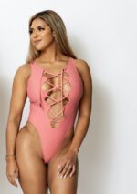 A blonde tanned model standing in front of a white wall wearing a pink lace up one piece swimsuit by vanity couture