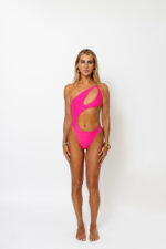 A blonde model standing in a. white studio wearing a hot pink asymmetrical one piece swimsuit with a gold chain strap by Vanity Couture.